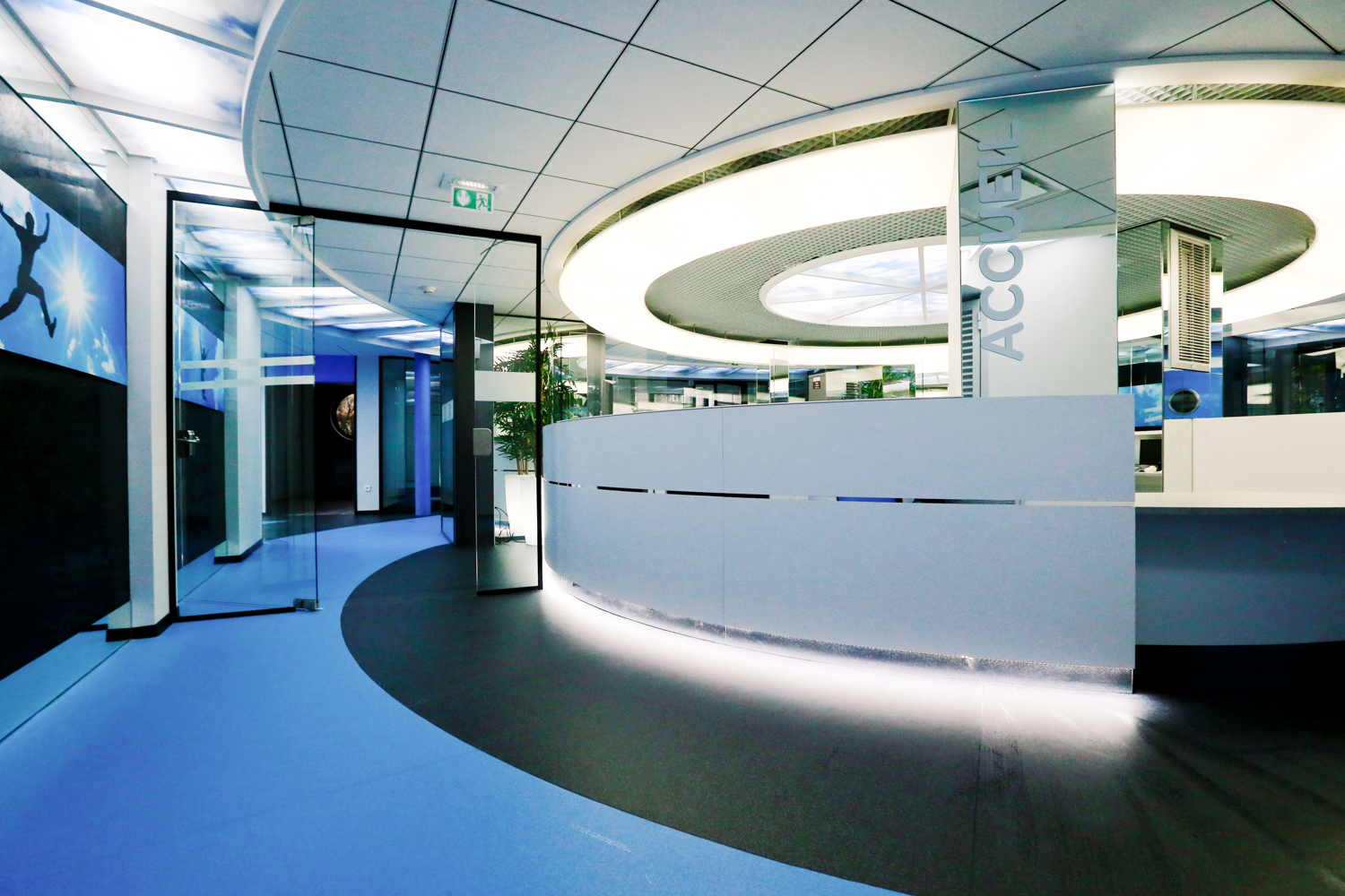 Espace accueil circulaire conçu par Arche du Bois, avec un miroir mural en face, une porte vitrée sur la gauche, et un luminaire LED circulaire au plafond diffusant des teintes bleues et blanches — un agencement immersif et contemporain.