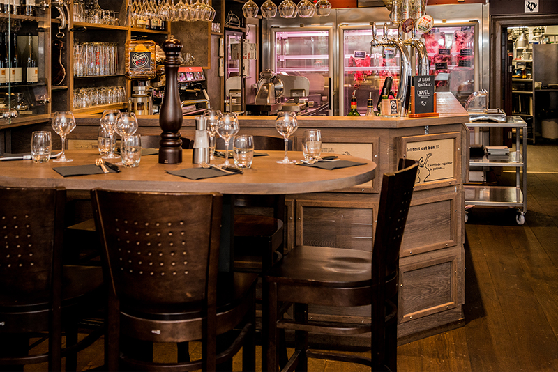 Table et chaises en bois avec des verres en verre posés dessus, placées devant un comptoir de bar en bois, lui-même adossé à un congélateur visible en arrière-plan — un agencement convivial et pratique signé Arche du Bois, mêlant authenticité et fonctionnalité.