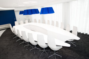 Table design à l’ergonomie originale entourée de chaises blanches, surmontée de suspensions bleues modernes ; en arrière-plan, une grande fenêtre habillée de rideaux Fabricant de mobilier sur mesure en Alsace