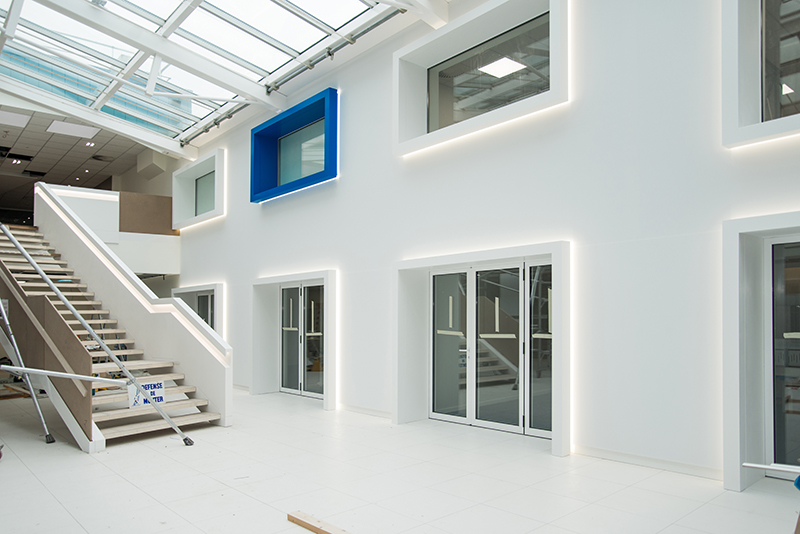 Vue d’ensemble d’un grand hall avec un escalier sur la gauche et un mur en face comportant plusieurs portes et fenêtres