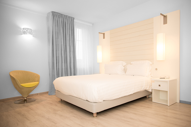 Lit double avec tête de lit en bois blanc, encadré par deux luminaires cylindriques suspendus au-dessus, et un fauteuil confortable placé sur la gauche de l’image — un agencement sur mesure signé Arche du Bois, alliant douceur, fonctionnalité et design contemporain.