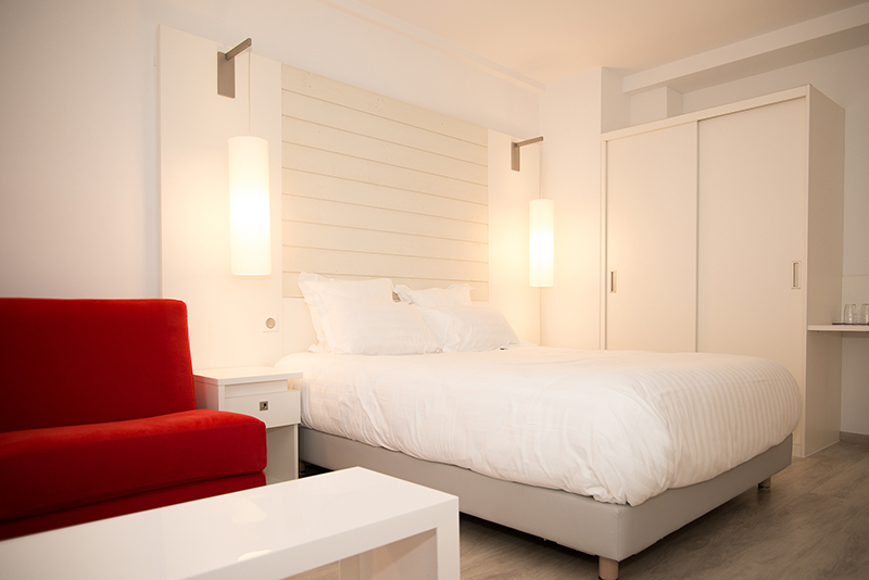 Lit blanc placé au centre de l’image, accompagné d’une armoire blanche sur le côté et de deux luminaires cylindriques fixés au-dessus de la tête de lit ; à gauche, un canapé rouge apporte une touche de couleur vive — un agencement harmonieux et contemporain signé Arche du Bois.