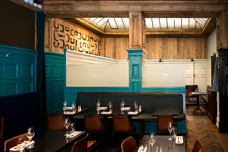 Intérieur de restaurant avec des tables et chaises de salle à manger, murs peints en bleu et plafond en bois apparent — un agencement chaleureux et contrasté signé Arche du Bois, alliant couleurs profondes et matériaux naturels.