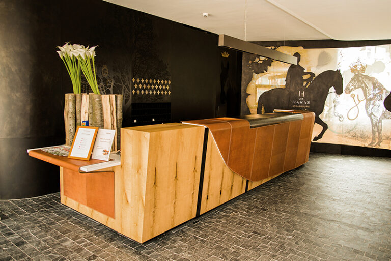 Comptoir d’accueil en bois avec un mur arrière noir apportant du contraste, et une fresque représentant deux chevaux sur le mur latéral droit — un agencement sur mesure signé Arche du Bois, mêlant élégance naturelle et expression artistique.