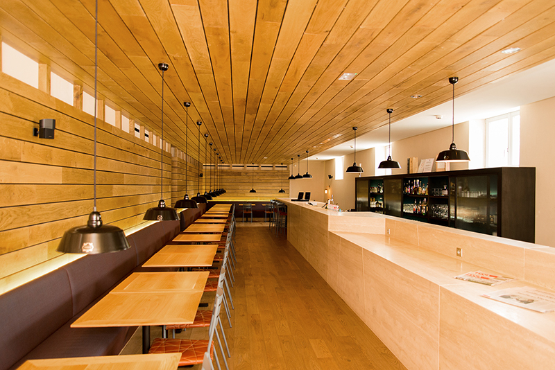 À gauche de l’image, plusieurs tables et chaises de salle à manger ; à droite, un comptoir en bois courant sur toute la longueur, avec des suspensions lumineuses au-dessus des tables ; l’ensemble de l’espace est habillé de bois au niveau du plafond et des murs — un agencement chaleureux et authentique réalisé par Arche du Bois.