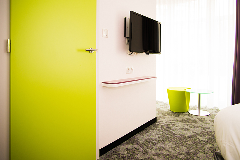 Vue intérieure d’une pièce aux murs verts, avec un téléviseur fixé au mur et un sol recouvert de moquette
