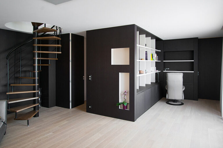 Salle contemporaine conçue par Arche du Bois, avec des murs noirs élégants, un escalier en colimaçon à gauche, et en face un ensemble de rangement mural comprenant une étagère intégrée et un placard bois sur mesure