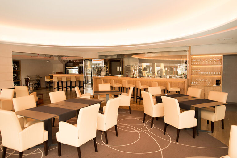 Salle à manger avec tables en bois foncé et chaises blanches disposées harmonieusement, avec un comptoir de bar en arrière-plan — un agencement contrasté et raffiné signé Arche du Bois, alliant convivialité et élégance dans un espace de restauration chaleureux.