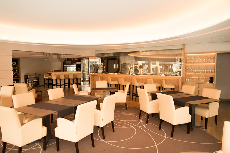 Salle à manger avec tables en bois foncé et chaises blanches disposées harmonieusement, avec un comptoir de bar en arrière-plan — un agencement contrasté et raffiné signé Arche du Bois, alliant convivialité et élégance dans un espace de restauration chaleureux.
