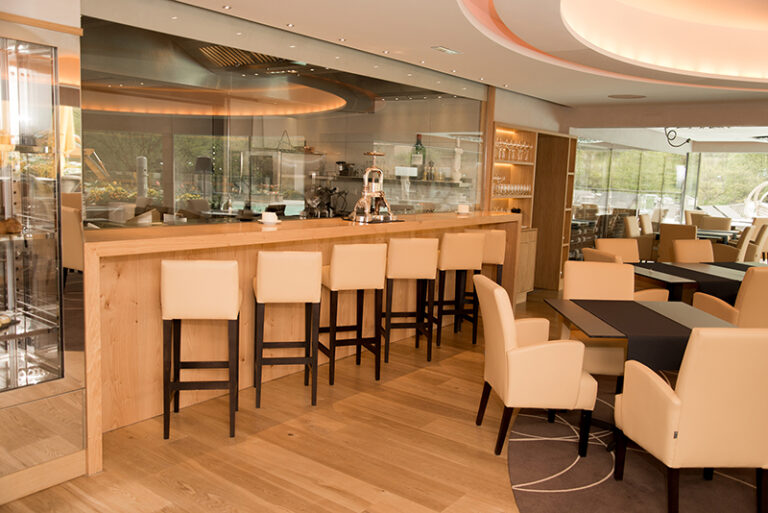 Bar avec comptoir en bois et rangée de tabourets alignés à l’avant, adossé à une cloison vitrée en fond — un agencement convivial et lumineux signé Arche du Bois, combinant mobilier sur mesure et transparence pour un espace de détente raffiné.