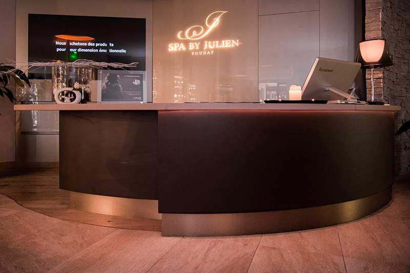 Comptoir d’accueil en bois de forme incurvée avec l’inscription lumineuse "Spa by Julien" à l’arrière
