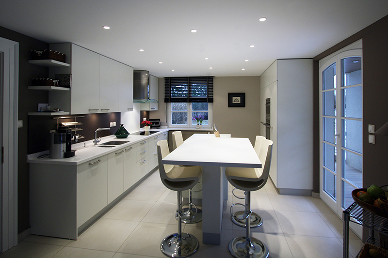Cuisine sur mesure par LDC Agencement – L’Arche du Bois : table centrale avec quatre tabourets, placards hauts et bas à gauche avec plaque de cuisson, lavabo et plan de travail, fenêtre au fond, et à droite placards, réfrigérateur et porte vitrée.