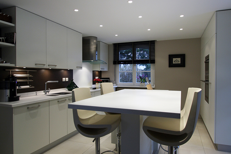 Cuisine sur mesure par LDC Agencement – L’Arche du Bois : table centrale avec quatre tabourets, placards hauts et bas à gauche avec plaque de cuisson, lavabo et plan de travail, fenêtre au fond, et à droite placards, réfrigérateur.