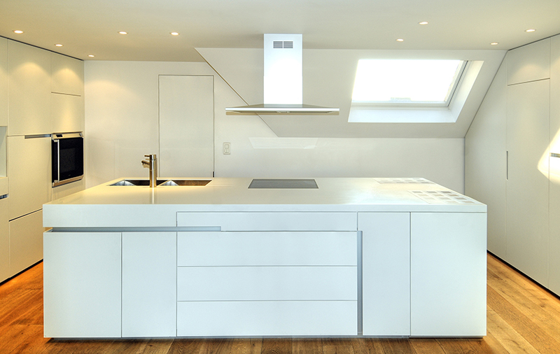 Cuisine sur mesure par LDC Agencement – L’Arche du Bois : îlot central blanc avec lavabo et plaque de cuisson intégrés, tiroirs intégrés, placards et étagères à gauche et fenêtre avec hotte au fond.