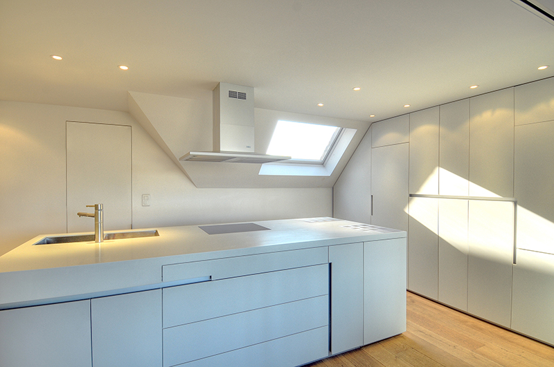 Cuisine sur mesure par LDC Agencement – L’Arche du Bois : îlot central blanc avec lavabo et plaque de cuisson intégrés, tiroirs intégrés, placards blancs à droite, et fenêtre avec hotte au fond.