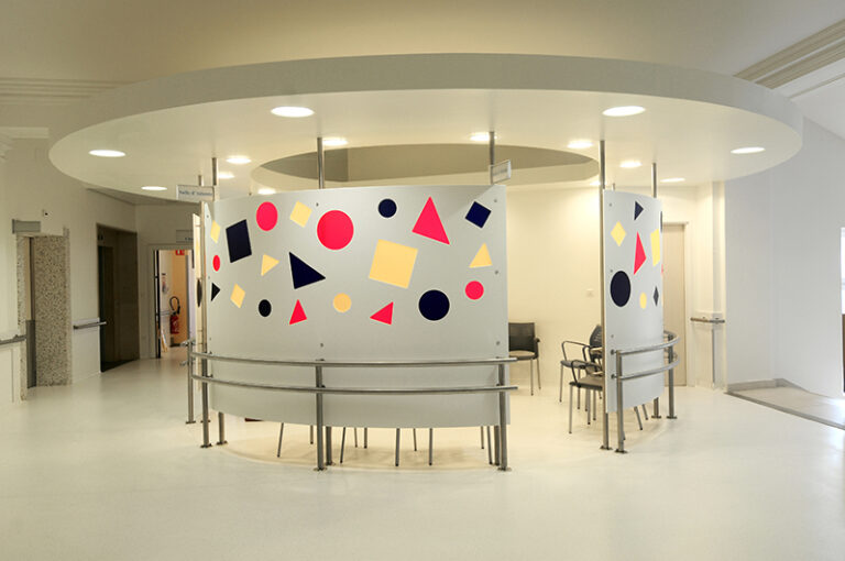Espace d’attente circulaire conçu par Arche du Bois, avec des formes géométriques (carrés, rectangles et triangles) dessinées sur les murs, surmonté d’une structure en arc intégrant un éclairage LED discret — un agencement à la fois artistique et fonctionnel.