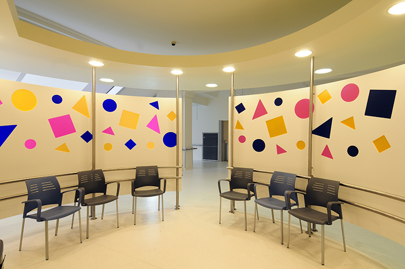 Espace d’attente circulaire conçu par Arche du Bois, avec des formes géométriques (carrés, rectangles et triangles) dessinées sur les murs, surmonté d’une structure en arc intégrant un éclairage LED discret — un agencement à la fois artistique et fonctionnel.