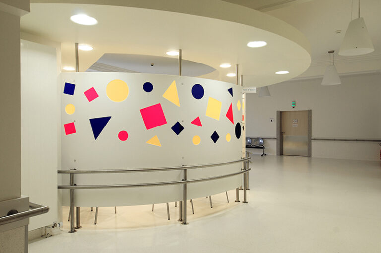 Espace d’attente circulaire conçu par Arche du Bois, avec des formes géométriques (carrés, rectangles et triangles) dessinées sur les murs, surmonté d’une structure en arc intégrant un éclairage LED discret — un agencement à la fois artistique et fonctionnel.