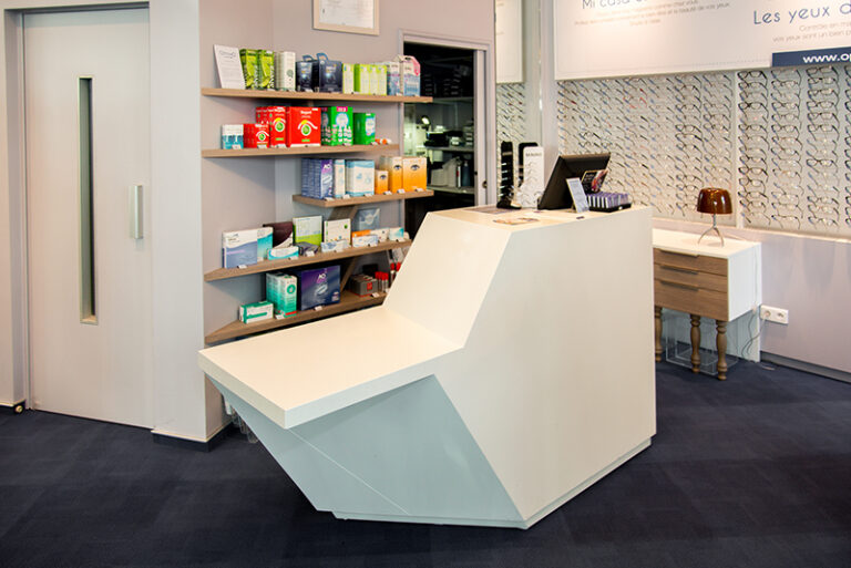 Comptoir d’accueil dans une boutique d’optique, avec à l’arrière des étagères contenant des lentilles de contact, et sur la droite des montures de lunettes de vue — un agencement fonctionnel et soigné réalisé par Arche du Bois.