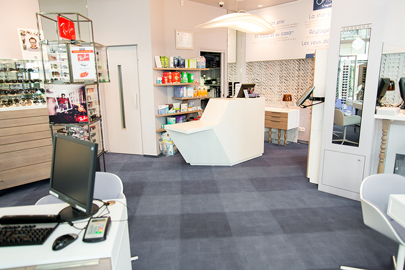 Intérieur de boutique d’optique avec un comptoir central et un sol recouvert de moquette — un agencement sur mesure réalisé par Arche du Bois, alliant confort, circulation fluide et esthétique professionnelle.