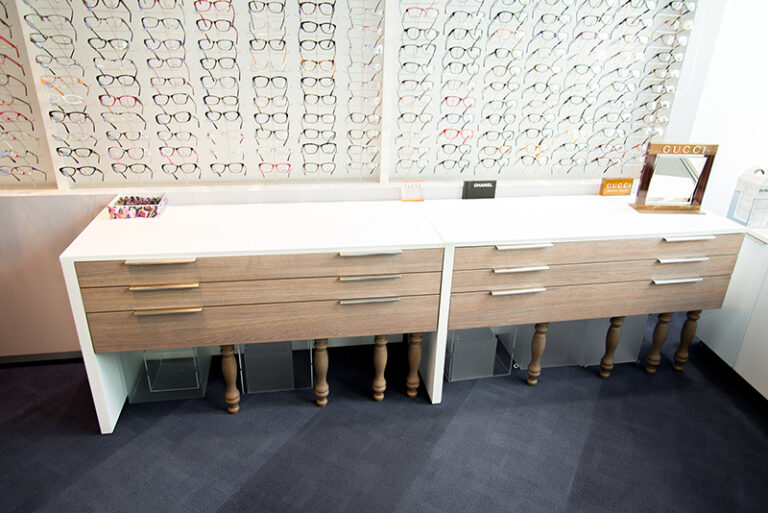 Étagères murales exposant des lunettes de vue, surmontant deux tables à tiroirs intégrés avec finition bois — un agencement sur mesure conçu par Arche du Bois, alliant présentation produit et fonctionnalité.