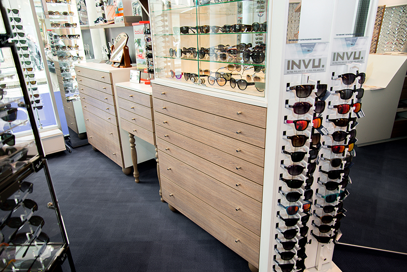 Étagères murales exposant des lunettes de vue, surmontant deux tables à tiroirs intégrés avec finition bois — un agencement sur mesure conçu par Arche du Bois, alliant présentation produit et fonctionnalité.