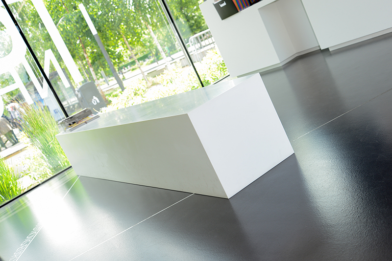 Grand bureau blanc placé au centre de l’image, avec un mur vitré à l’arrière laissant passer la lumière