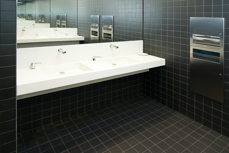 Deux lavabos blancs alignés devant un grand miroir mural, avec murs et sol recouverts de carrelage noir, et une petite poubelle visible sur la droite — un aménagement raffiné et contemporain signé Arche du Bois, jouant sur les contrastes de couleurs et les matériaux nobles.