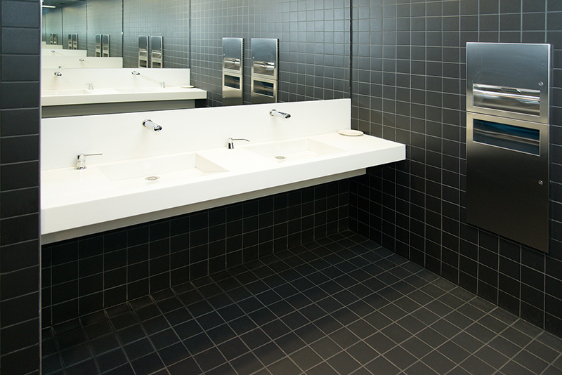 Deux lavabos blancs alignés devant un grand miroir mural, avec murs et sol recouverts de carrelage noir, et une petite poubelle visible sur la droite — un aménagement raffiné et contemporain signé Arche du Bois, jouant sur les contrastes de couleurs et les matériaux nobles.