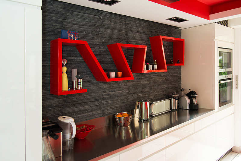 Étagère murale rouge dans une cuisine, installée au-dessus d’une table sur laquelle se trouvent divers appareils électroménagers tels qu’une machine à café, un grille-pain et d’autres ustensiles de cuisine. À droite de l’image, un four encastré complète l’espace fonctionnel