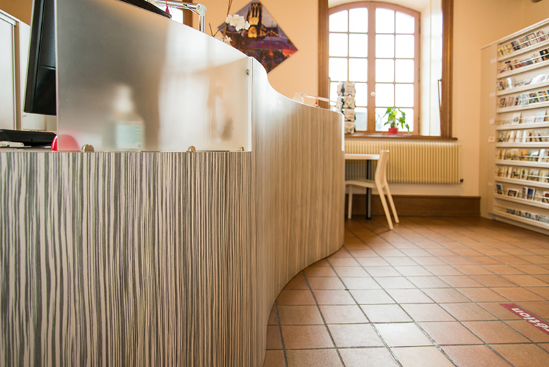 À gauche, un comptoir d’accueil en bois ; à droite, une étagère de rangement ; en face, une fenêtre avec un radiateur et un fauteuil placé devant