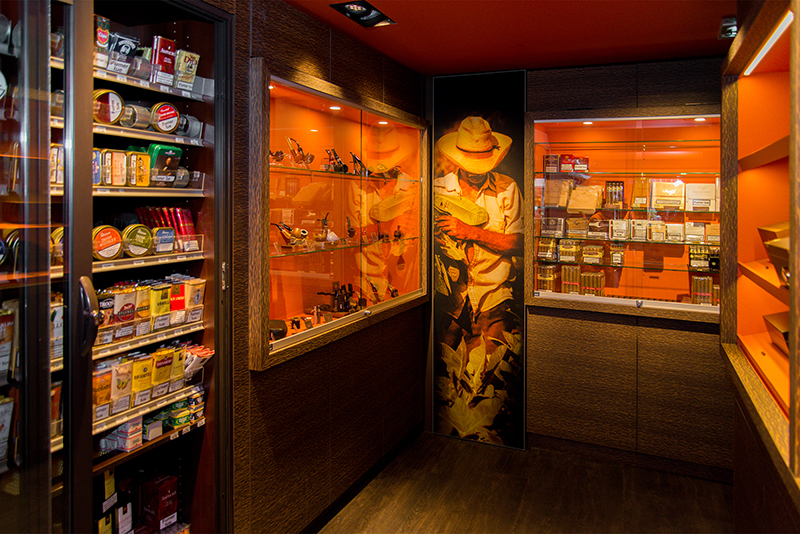 À gauche de l’image, une vitrine en verre exposant des pipes, tandis qu’en face se trouvent des paquets de cigarettes — un agencement raffiné signé Arche du Bois, alliant présentation produit et élégance du mobilier.