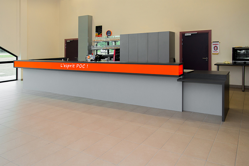 Comptoir de bar gris surmonté d’une bande orange distinctive, avec à l’arrière un placard intégré et une porte