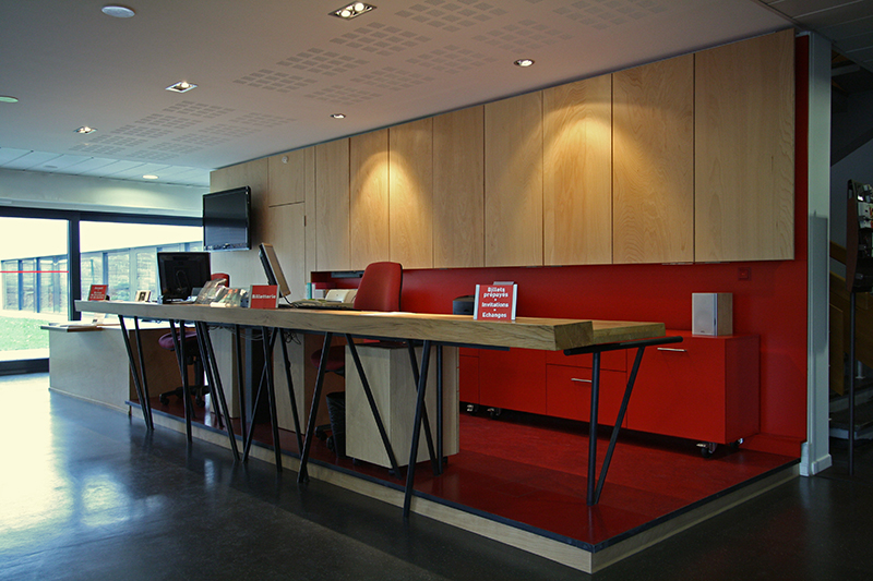 Comptoir d’accueil en bois avec piètement en métal noir, accompagné de rangements rouges positionnés à l’arrière sur le sol, surmontés de murs habillés en bois — une réalisation sur mesure par Arche du Bois, mêlant chaleur du bois, touches industrielles et accents de couleur pour un espace d’accueil distinctif
