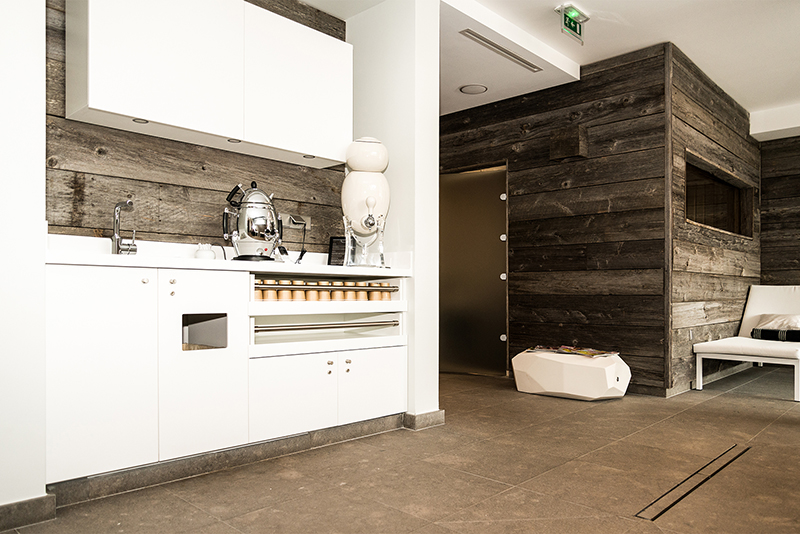 En face, une petite pièce au revêtement extérieur en bois naturel ; sur la gauche, un meuble bas et haut de cuisine blanc avec un samovar posé dessus — un agencement chaleureux et pratique signé Arche du Bois, mêlant tradition et fonctionnalité.