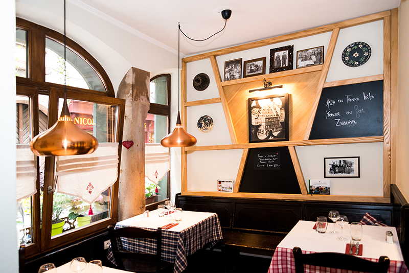 Deux tables accompagnées de chaises au centre de l’image, devant un mur habillé d’éléments décoratifs en bois, avec une fenêtre visible sur la gauche — un agencement harmonieux et lumineux signé Arche du Bois.