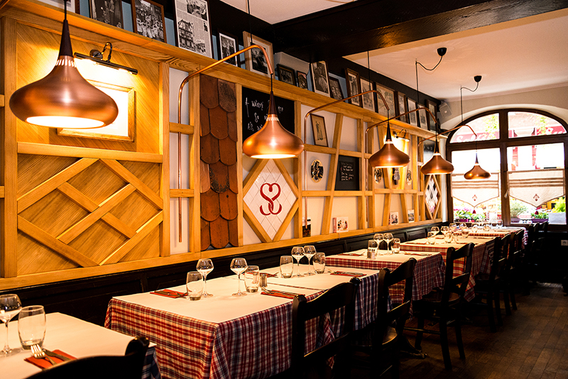 À gauche de l’image, des tables recouvertes de nappes accompagnées de chaises, devant un mur décoré d’éléments en bois avec des étagères intégrées contenant des objets, surmonté de plusieurs cadres accrochés — un agencement soigné et chaleureux signé Arche du Bois.