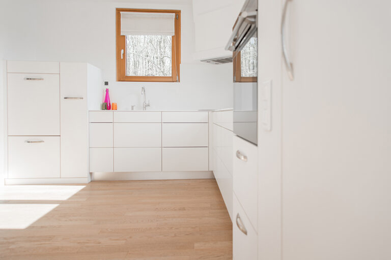 Vue d’une cuisine avec agencement en L, comprenant des armoires sur le mur de droite et en face, ainsi qu’une fenêtre intégrée dans le mur du fond pour un éclairage naturel