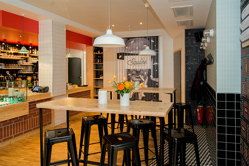 Deux tables hautes accompagnées de tabourets, placées dans une pièce dont les murs sont habillés de carreaux