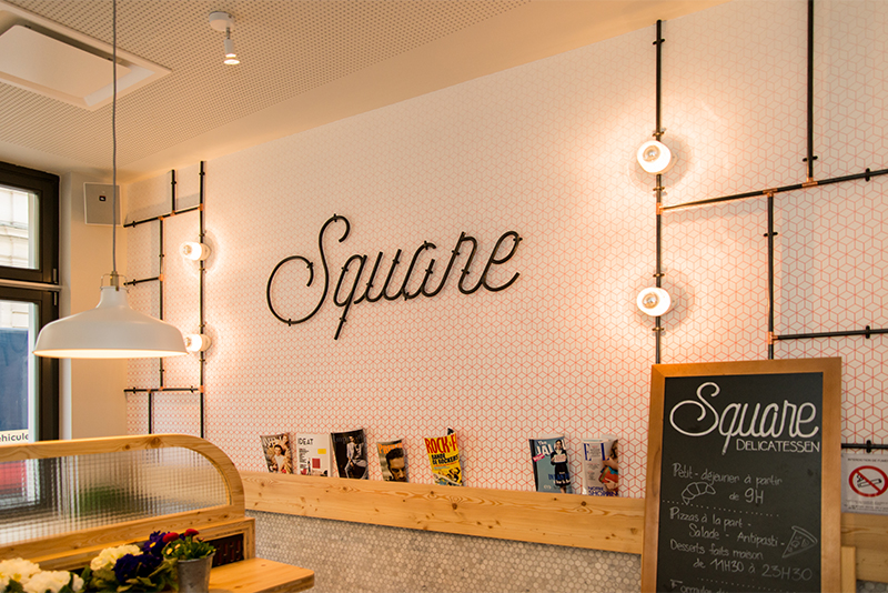 Mur décoratif avec le mot "Square" en lettres stylisées, accompagné de barres métalliques et d’un éclairage intégré pour une mise en valeur graphique ; un panneau noir est visible sur la droite — une composition contemporaine et structurée, réalisée par Arche du Bois.