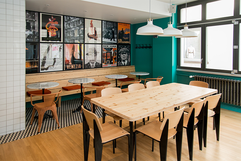 Grande table de salle à manger pour huit personnes, éclairée par trois suspensions alignées ; à l’arrière, une seconde table est visible, avec une fenêtre sur la droite et plusieurs cadres décoratifs accrochés au mur du fond — un agencement soigné et accueillant signé Arche du Bois.