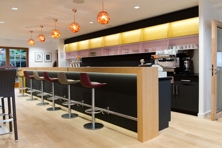 Comptoir de bar avec plateau en bois naturel et structure noire, accompagné de six tabourets alignés ; cinq suspensions lumineuses au-dessus et éclairage LED intégré en partie basse — un agencement convivial et moderne signé Arche du Bois.