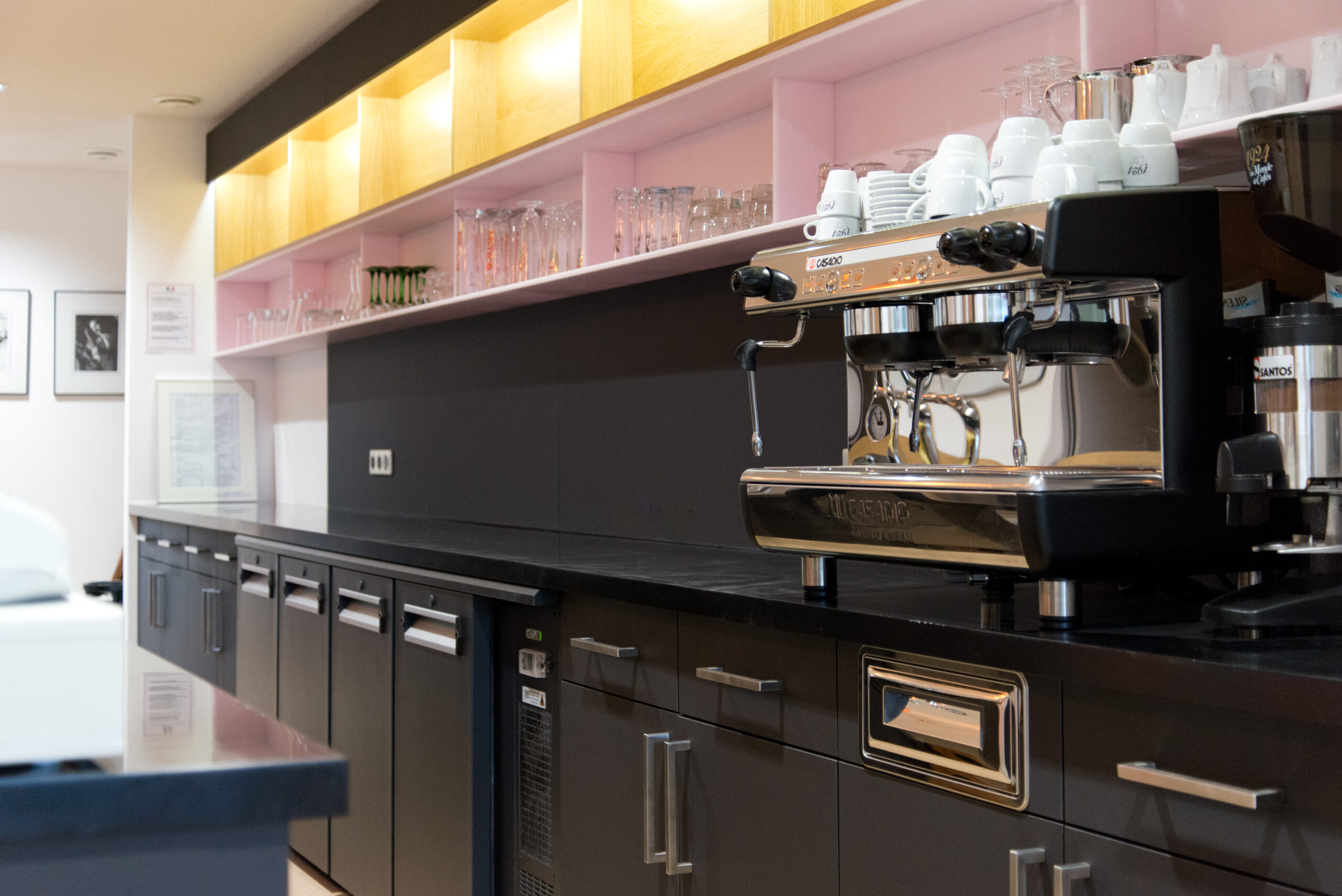 Vue rapprochée d’un comptoir de bar avec machine à café posée sur un plan en bois, meubles inférieurs noirs et étagères à verres rétroéclairées en hauteur — un agencement fonctionnel et raffiné signé Arche du Bois, pensé pour les espaces de restauration haut de gamme.