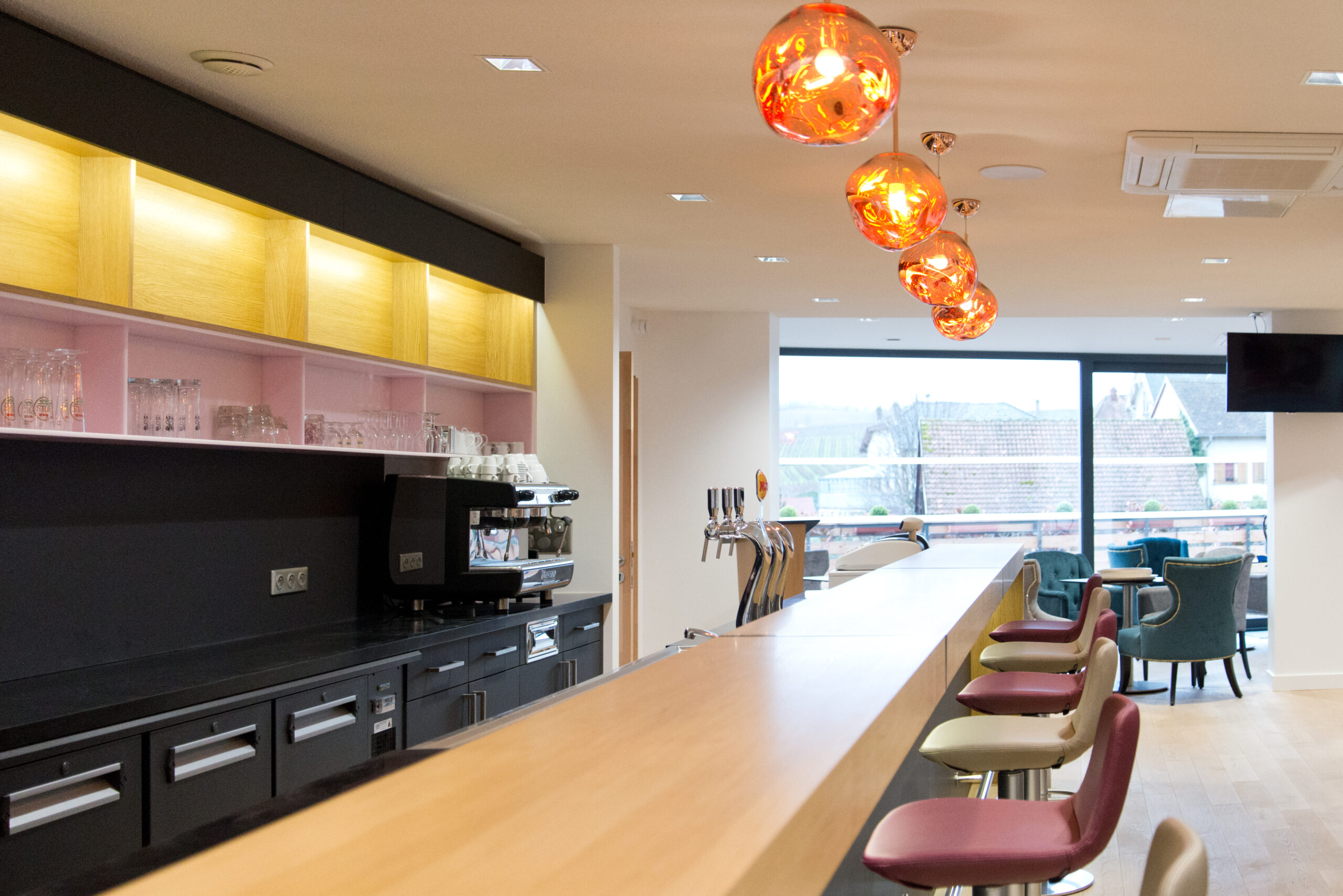 Vue depuis le dessus d’un comptoir de bar avec plusieurs tabourets alignés à l’arrière, et une grande baie vitrée en fond de pièce — un agencement lumineux et chaleureux signé Arche du Bois, combinant mobilier sur mesure et ouverture vers l’extérieur.