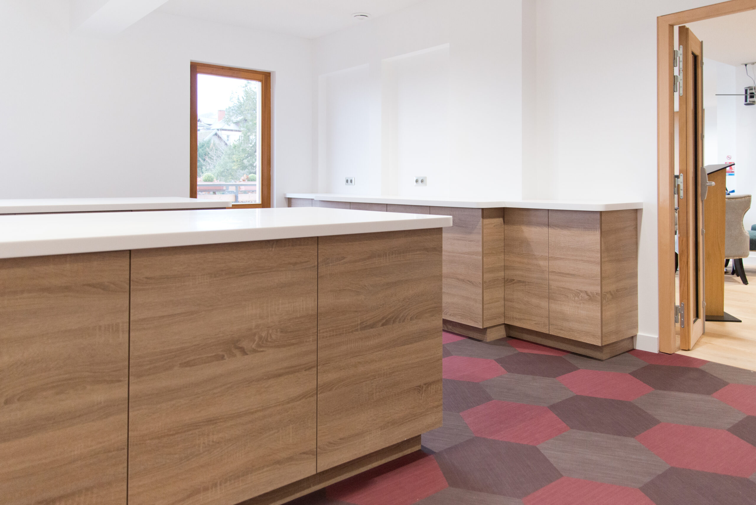 Ensemble de meubles bas en bois avec plans de travail blancs, encadrés par une porte à droite et une fenêtre à gauche