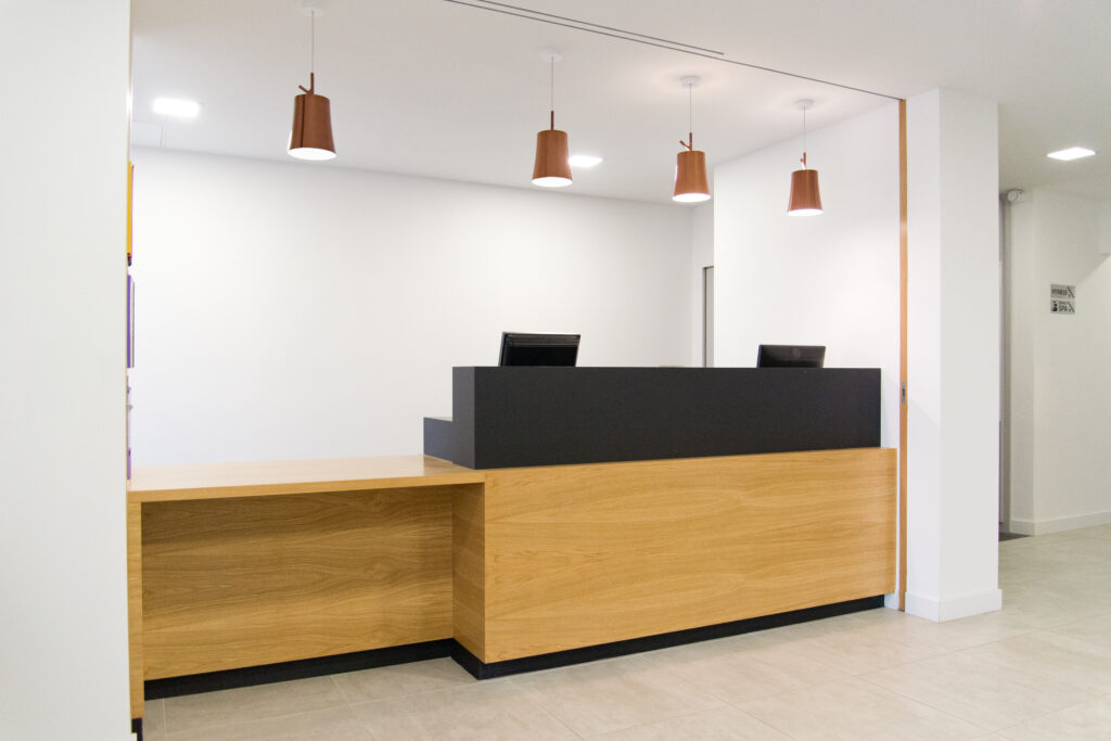 Comptoir d’accueil en bois avec une partie supérieure noire, surmonté de quatre suspensions lumineuses, entouré de murs blancs — un agencement moderne et contrasté signé Arche du Bois, alliant chaleur du bois et sobriété des teintes.