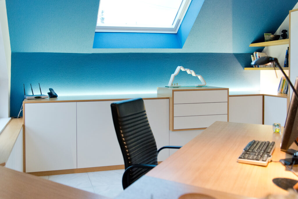 Bureau en bois avec une chaise noire, placé devant une armoire blanche encadrée de bois, sur fond de mur bleu. Un puits de lumière au plafond éclaire naturellement l’espace de travail conçu par Arche du Bois
