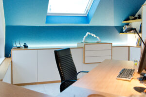 Bureau en bois avec une chaise noire, placé devant une armoire blanche encadrée de bois, sur fond de mur bleu. Un puits de lumière au plafond éclaire naturellement l’espace de travail conçu par Arche du Bois. Agencement Intérieur Particuliers