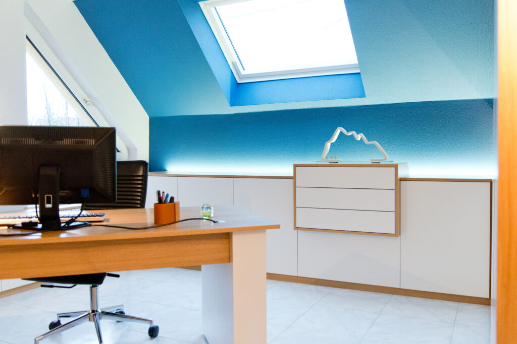 Bureau de travail avec écran posé sur un plateau en bois clair, adossé à une armoire blanche encadrée de bois, le tout devant un mur bleu avec fenêtre de toit apportant une lumière naturelle – aménagement réalisé par Arche du Bois.