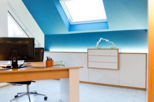 Bureau sur mesure Alsace avec écran posé sur un plateau en bois clair, adossé à une armoire blanche encadrée de bois, le tout devant un mur bleu avec fenêtre de toit apportant une lumière naturelle – aménagement réalisé par Arche du Bois.